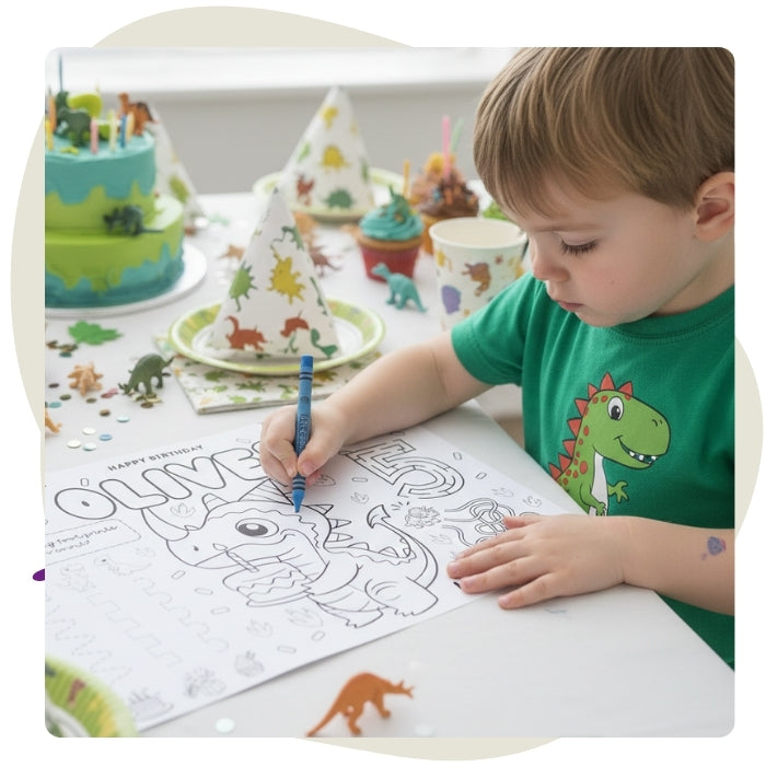 A top-down view of a dinosaur-themed birthday party table setup. The centerpiece is a personalized black-and-white coloring placemat for a child named Oliver turning five. The table is surrounded by colorful cupcakes, a two-tiered green and blue dinosaur cake, scattering of small plastic dinosaur toys, confetti, and dinosaur-patterned plates, cups, and party hats.